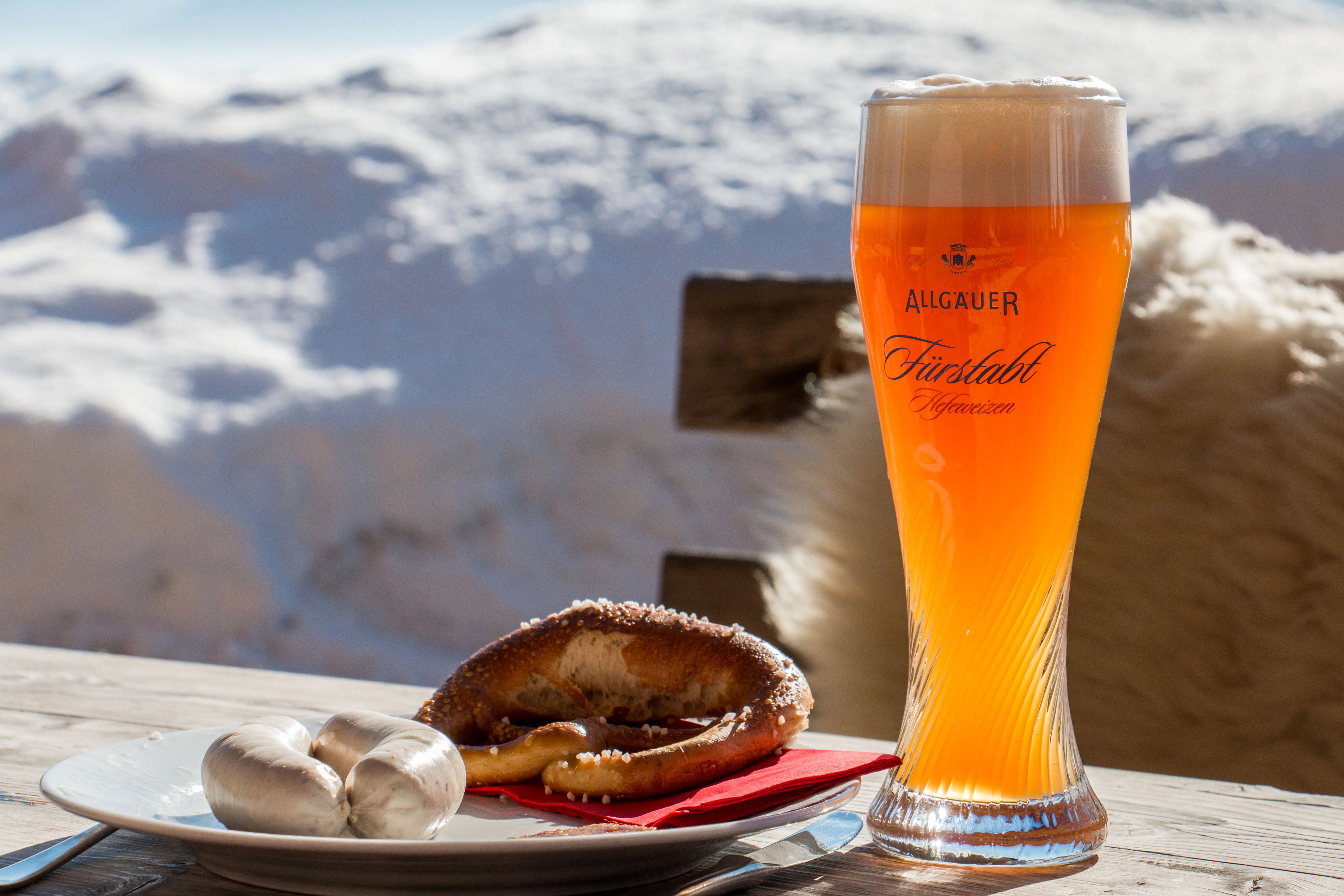 Ein Weißen, ein Teller wo zwei Weißwurste und eine Breze drauf sind, mit einem schönen Alpenausschnitt im Hintergrund.