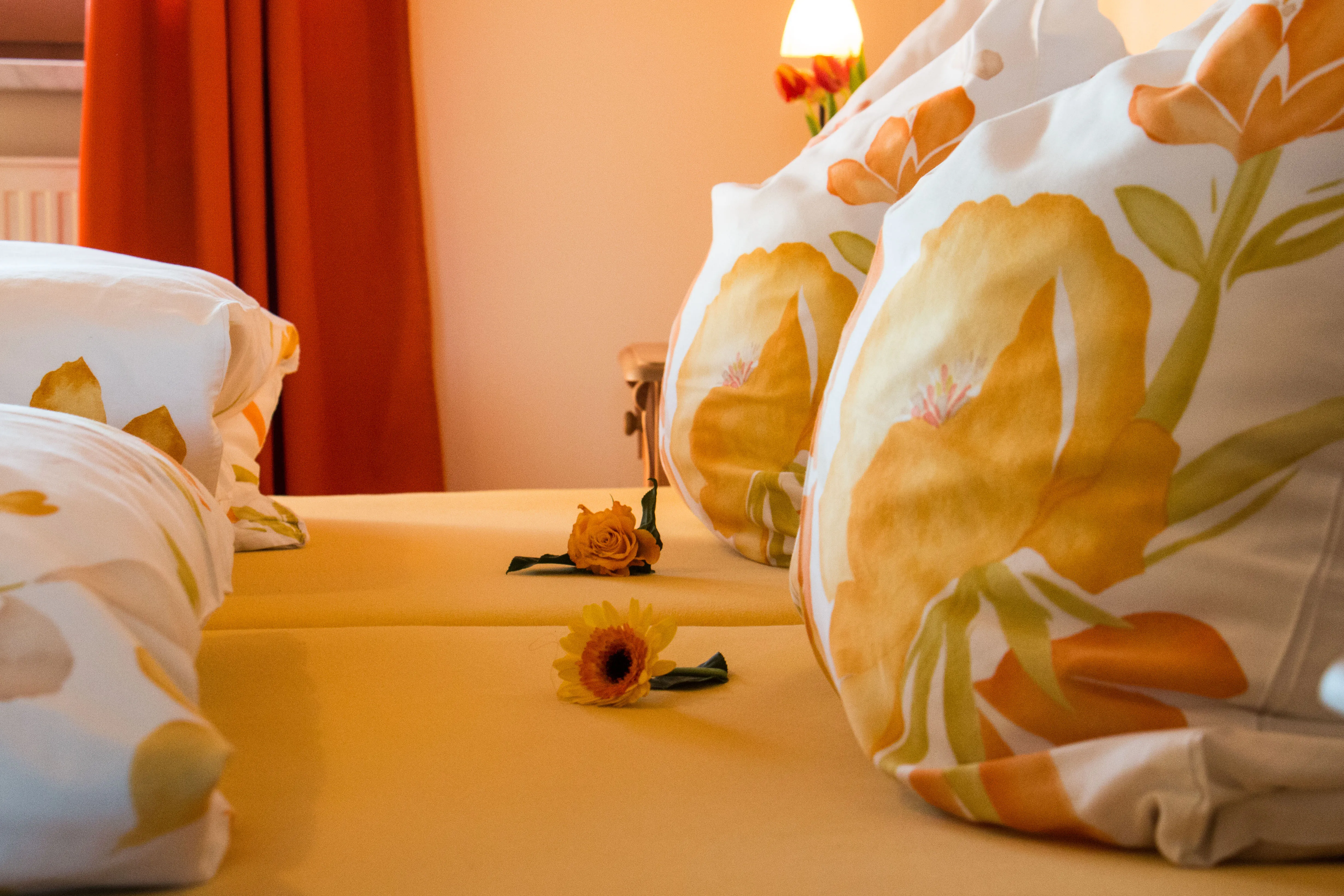 Nahaufnahme eines Gasthof Bettes mit 2 verschiedene Blumen zwischen den Kissen mit Blumenkissenbezug und den Decken auf gelb orangen Bettbezug.