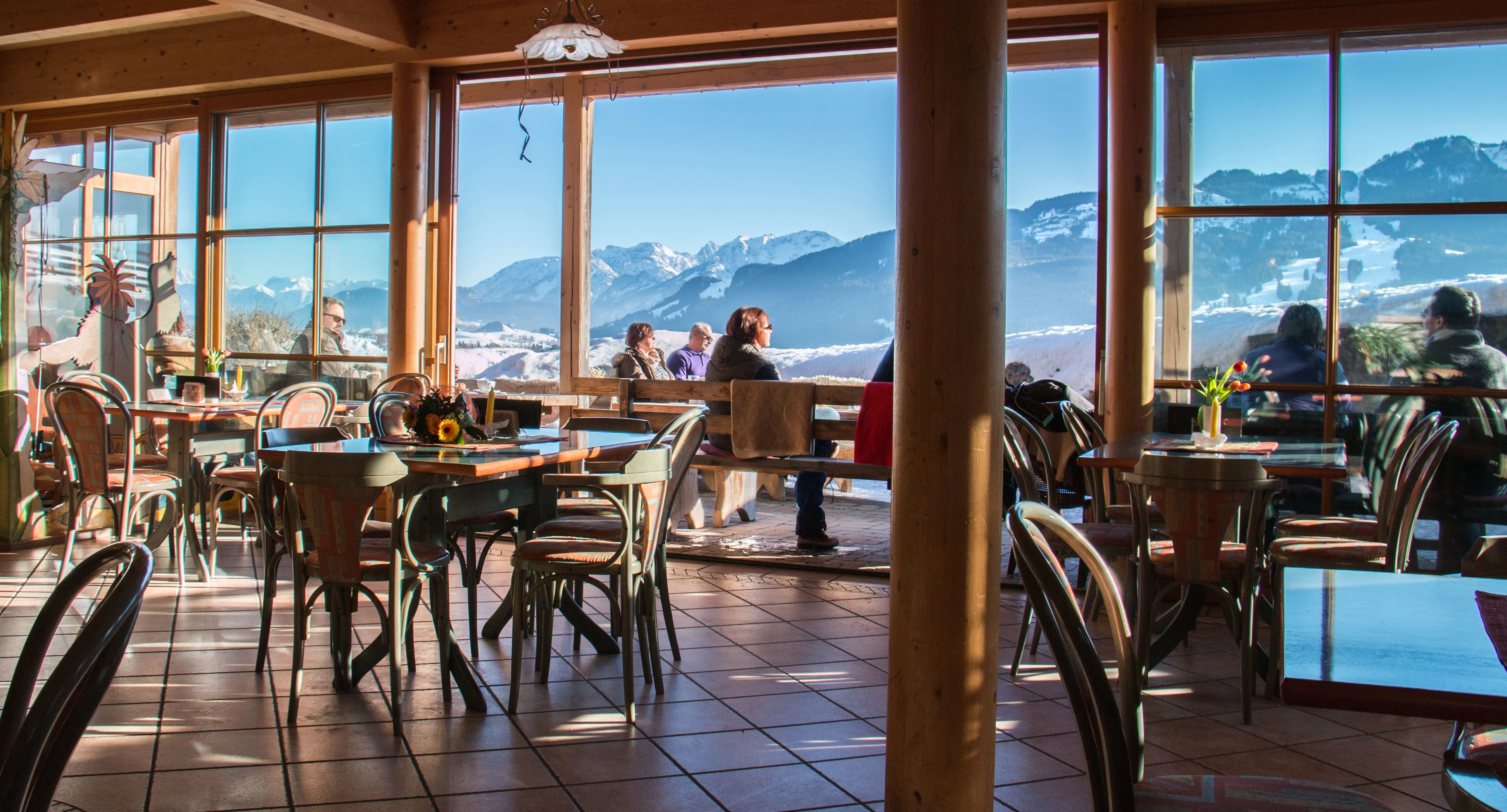 Aussicht aus dem innerem Gastraum, welches auf as Winter Alpenpanorama und verschiedene Menschen schaut.